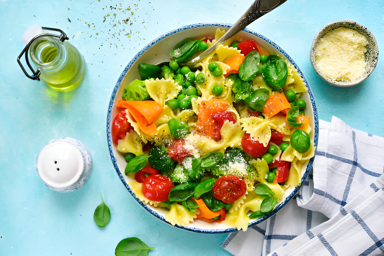 A vibrant bowl of pasta salad featuring farfalle pasta, cherry tomatoes, green peas, and fresh basil on a blue background.