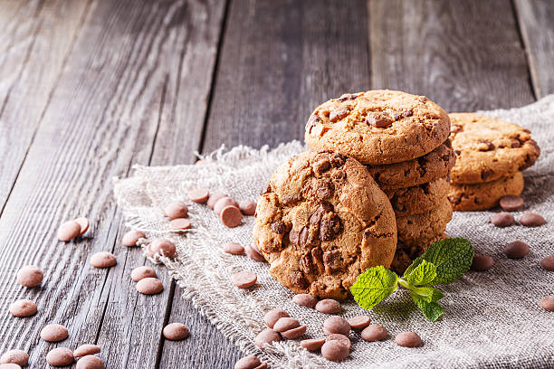 A stack of chocolate chip cookies on a burlap cloth with chocolate chips scattered around and fresh mint leaves.