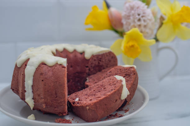 A sliced chocolate bundt cake with white icing served on a plate, accompanied by a bouquet of yellow and pink flowers in a white vase.
