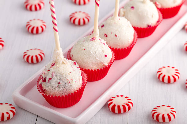 Five cake pops decorated with coconut and pink sprinkles, placed on a white platter with red and white striped candies scattered around.