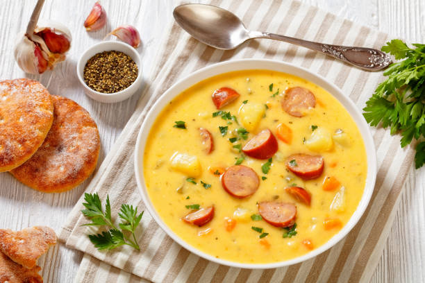 A bowl of creamy soup with chunks of sausage, vegetables, and potatoes, served with bread and garlic on the side.