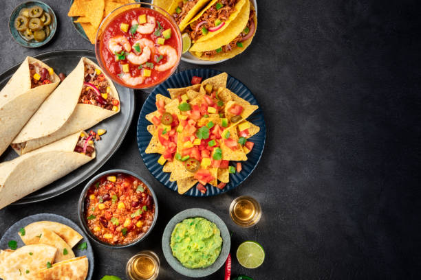 A variety of Mexican dishes including tacos, nachos, guacamole, and salsa on a dark background.