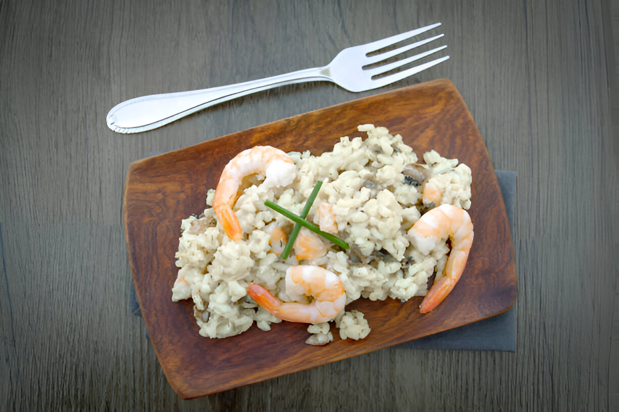 A wooden plate with creamy risotto topped with shrimp and chives, accompanied by a fork on a wooden table.