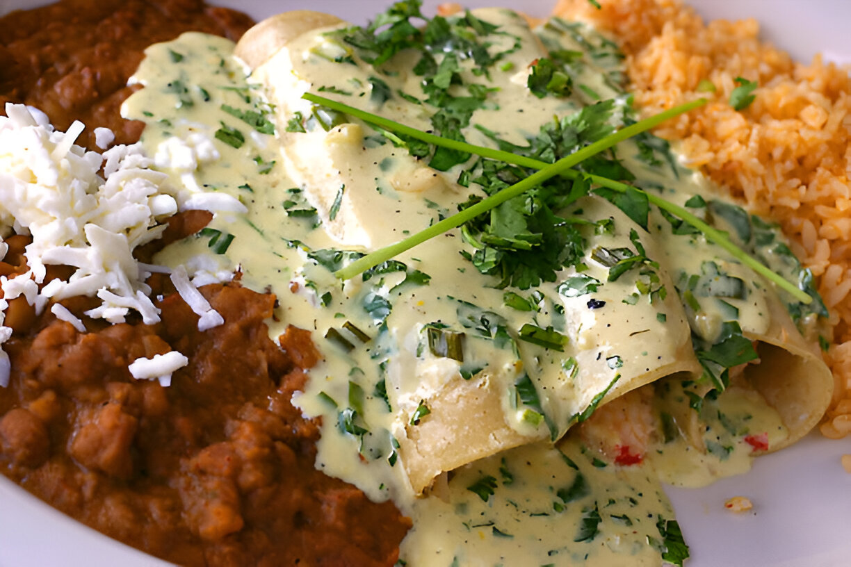 A plate of enchiladas topped with creamy green sauce and fresh herbs, served with refried beans and Spanish rice.