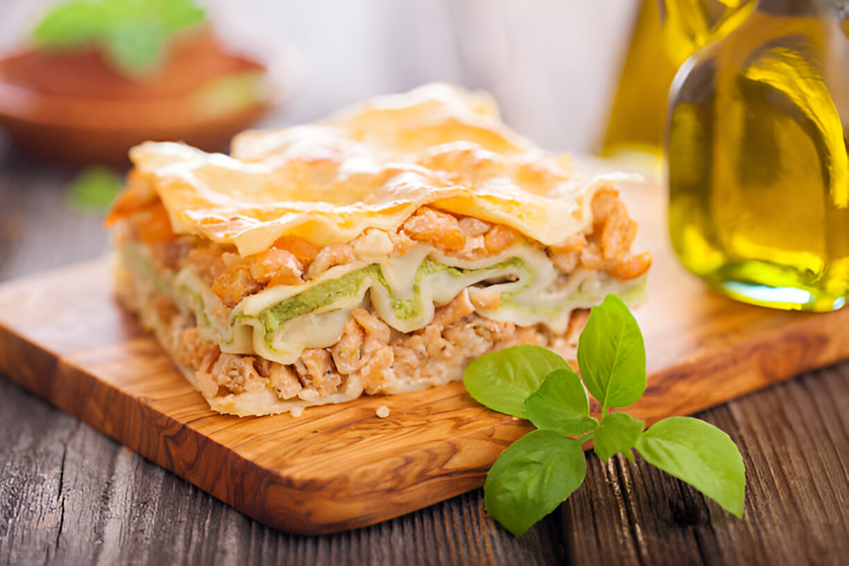 A delectable slice of lasagna with layers of green, white, and orange ingredients, served on a wooden cutting board with a basil leaf and olive oil bottles in the background.