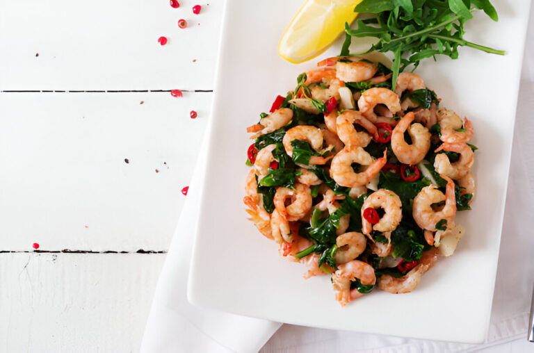 A plate of shrimp and greens with red chili and a lemon wedge, served on a white table.