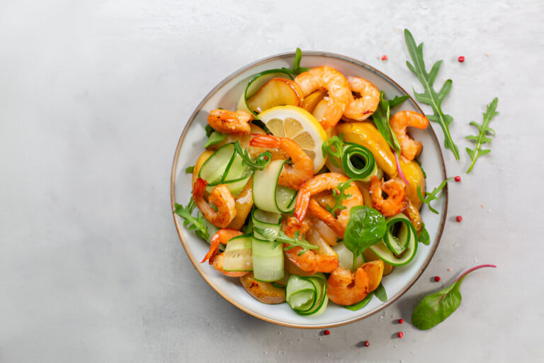 A vibrant salad featuring shrimp, cucumber slices, arugula, and lemon on a light gray background.