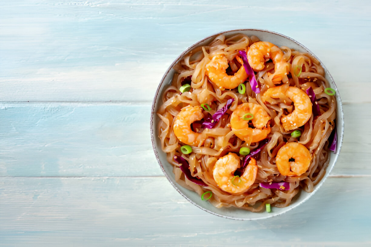 A bowl of shrimp noodles topped with purple cabbage and green onions on a light wooden surface.