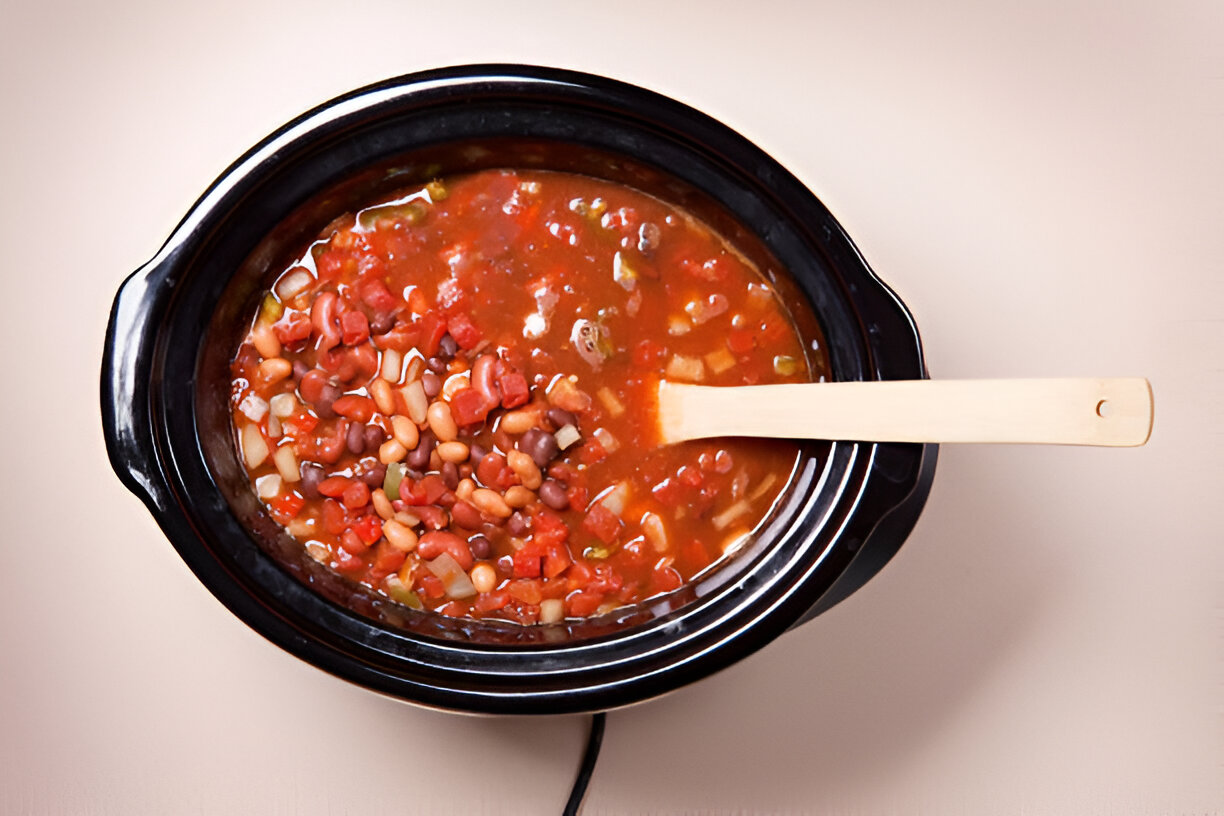 A slow cooker filled with a mixed bean stew featuring red and white beans, tomatoes, and vegetables, with a wooden spoon resting on the edge.