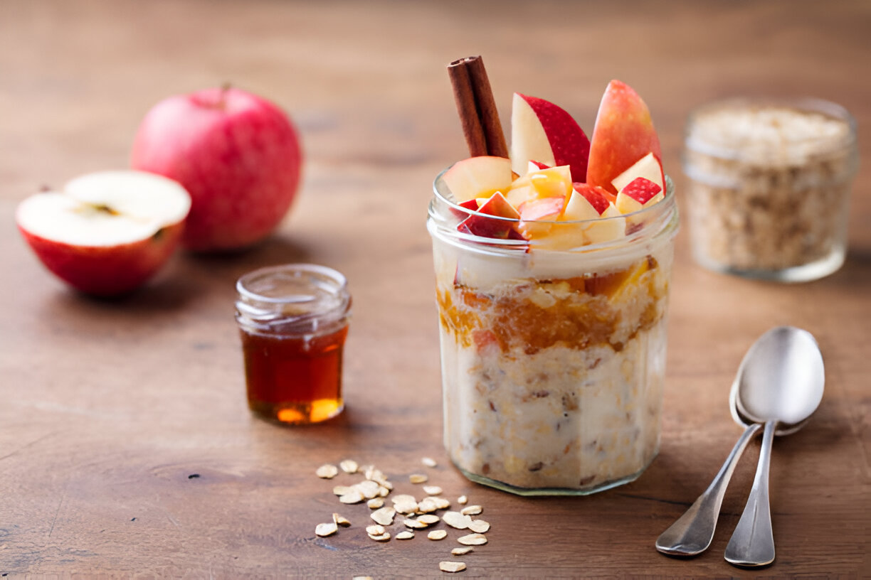 A jar of layered oatmeal dessert with apples, cinnamon, and honey, surrounded by ingredients like a whole apple, honey jar, and oats on a wooden surface.