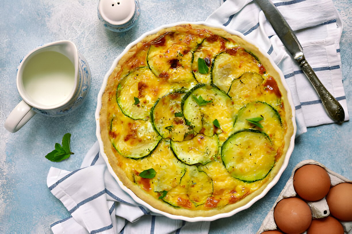 A delicious zucchini quiche in a white pie dish, garnished with fresh herbs, accompanied by eggs and a milk pitcher.