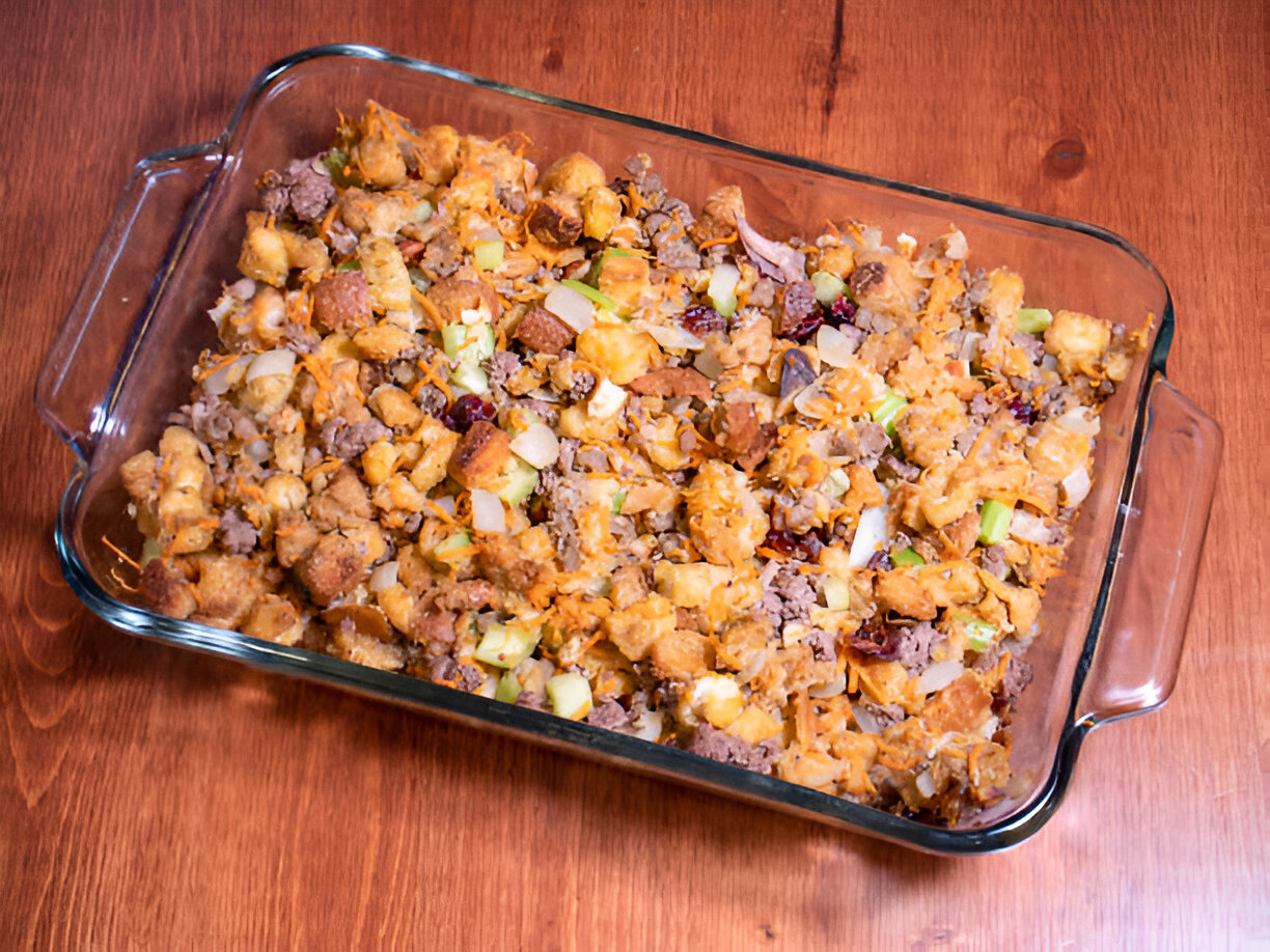 A glass baking dish filled with a savory stuffing mixture containing bread, ground meat, vegetables, and seasonings.