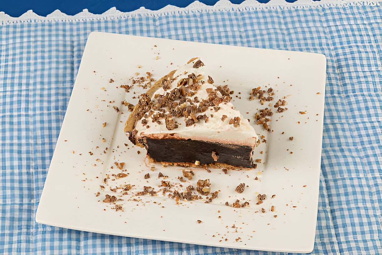 A slice of chocolate cream pie on a white plate with crushed nuts on top, placed on a blue checkered tablecloth.