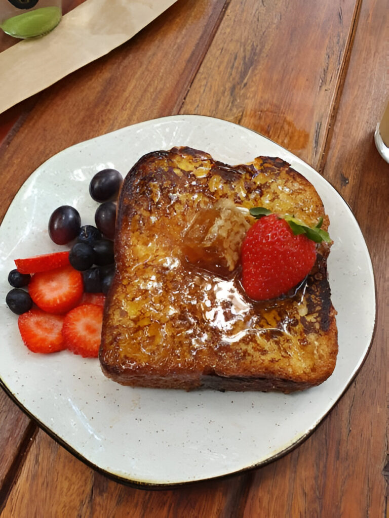 A plate of French toast topped with syrup and a sliced strawberry, accompanied by fresh berries on the side.