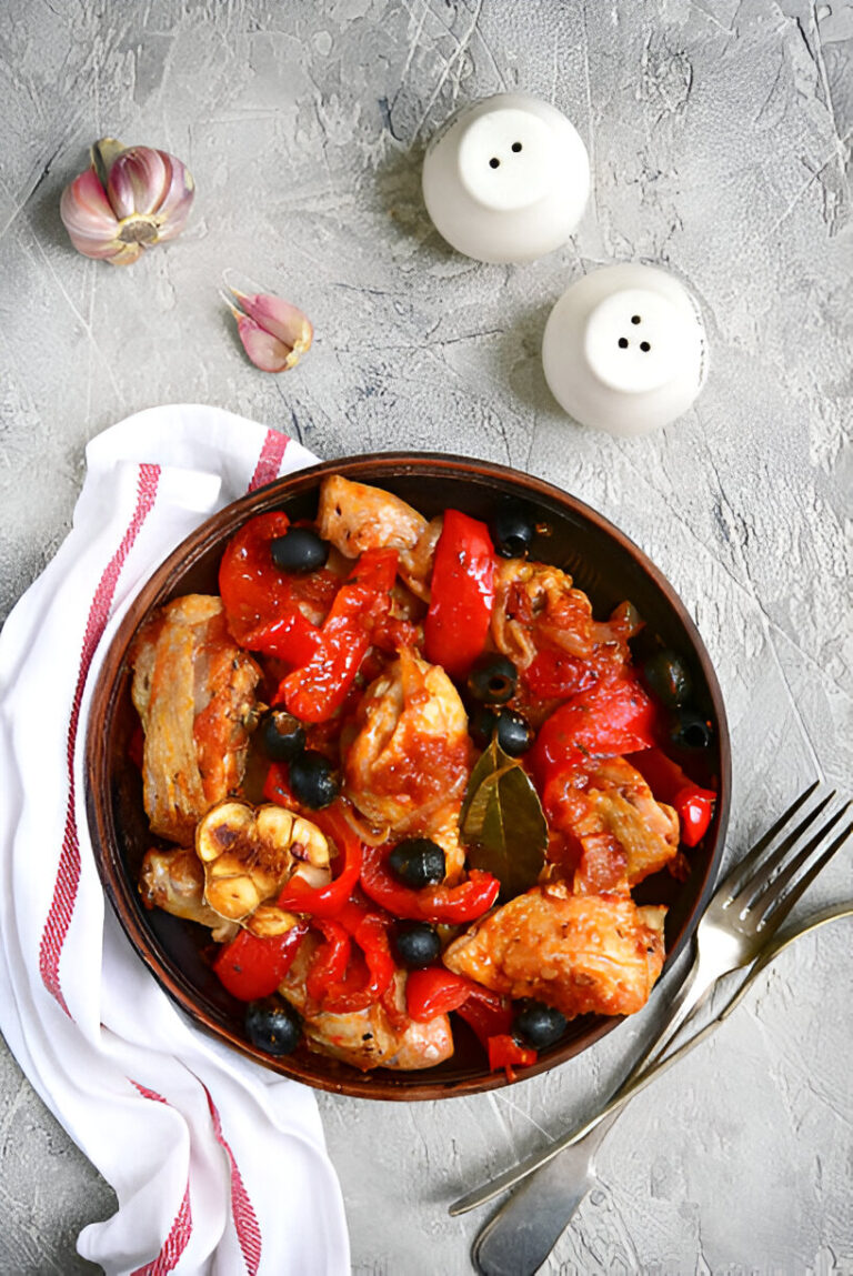 A bowl of chicken stew with red peppers, olives, and garlic on a textured gray surface.