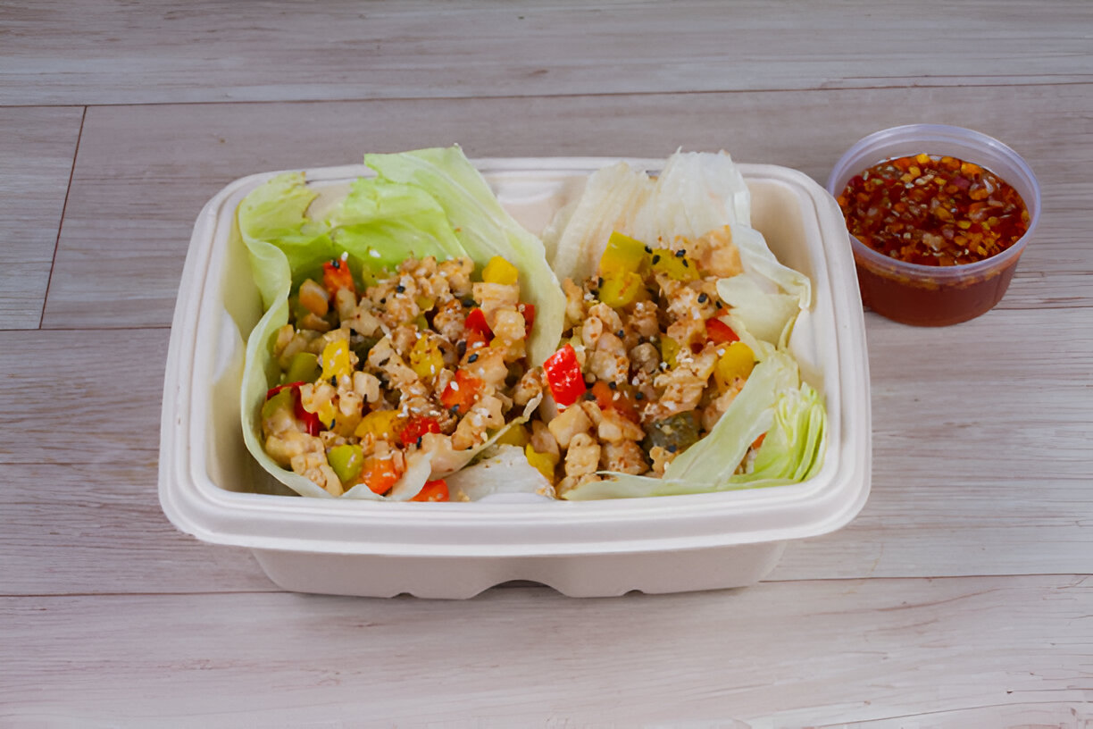 A fresh meal served in a biodegradable container, featuring two lettuce wraps filled with colorful vegetables and a side of dipping sauce.