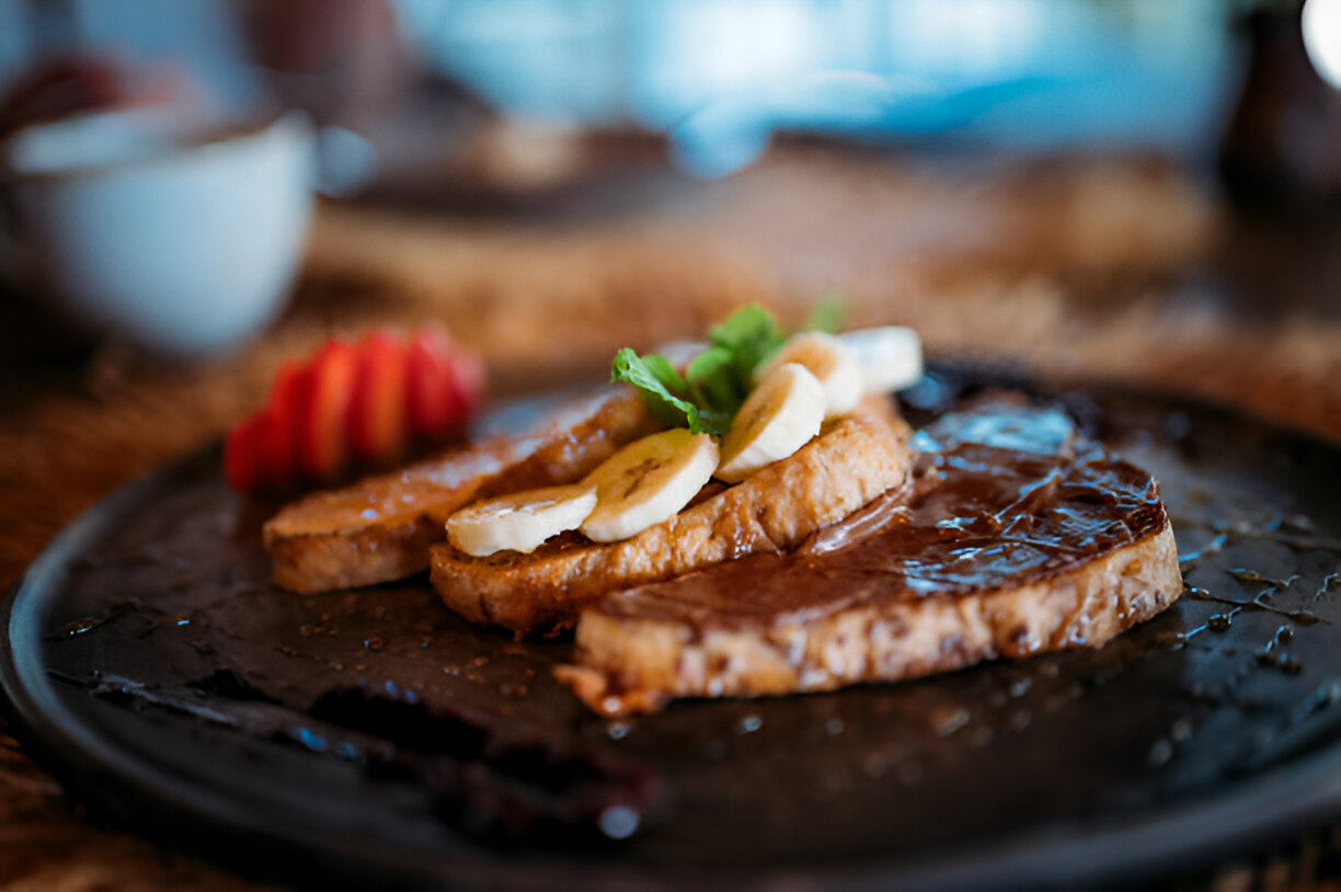 Delicious French toast topped with banana slices and a sprig of mint, served on a dark plate with strawberries in the background.