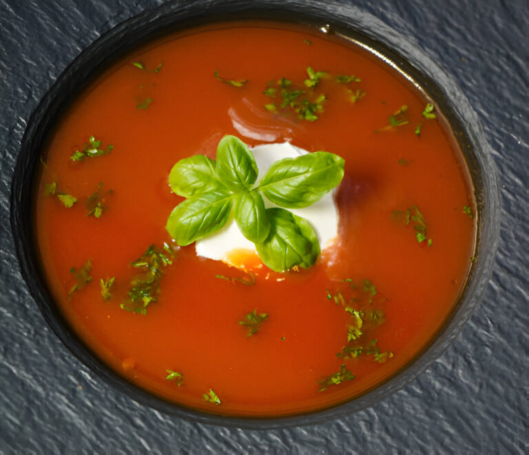 Tomato soup garnished with a basil leaf and fresh herbs in a dark bowl on a textured surface.
