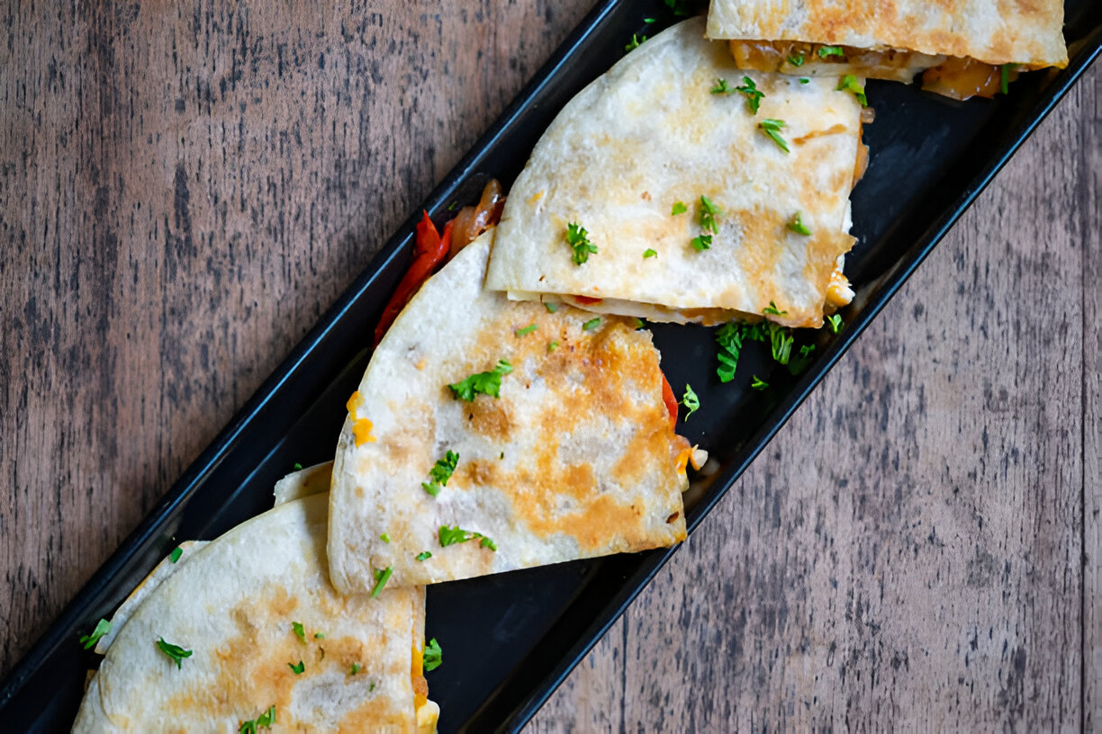 Four golden-brown quesadilla slices garnished with fresh parsley on a black plate.