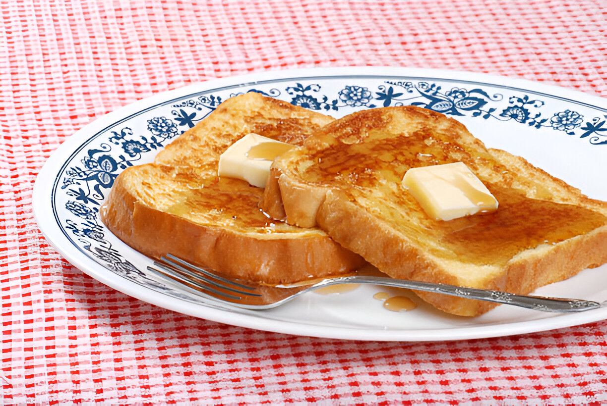 A plate of golden-brown French toast topped with butter and syrup, sitting on a red and white checkered tablecloth.