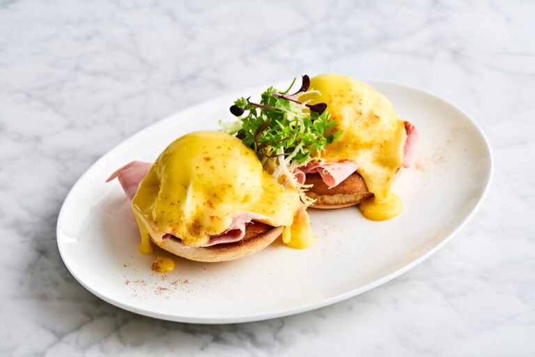 Two delicious eggs benedict on a white plate topped with hollandaise sauce and ham, garnished with fresh greens.
