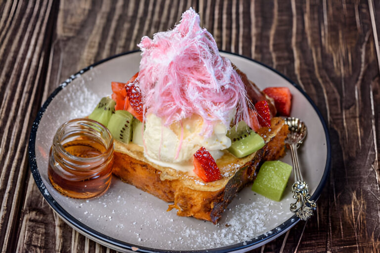 A decadent dessert featuring thick toast topped with ice cream, cotton candy, and assorted fruits, served with a small jar of syrup.