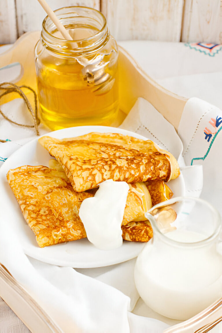 A plate of golden-brown crepes served with honey and yogurt, accompanied by a jar of honey and a small creamer of milk.