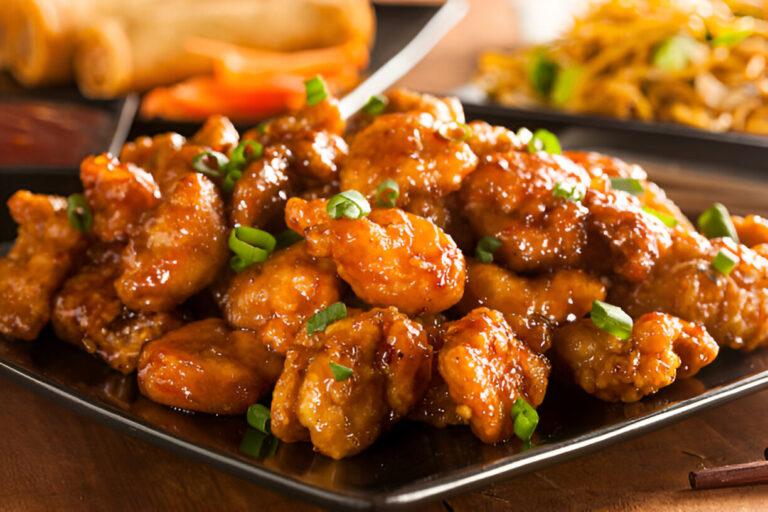 A close-up of a plate filled with crispy, glazed chicken pieces garnished with green onions, served with spring rolls in the background.