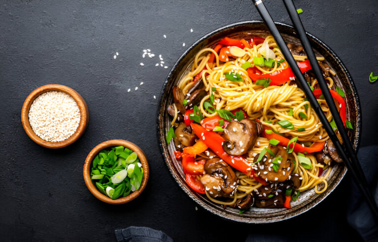 A bowl of stir-fried noodles with bell peppers, mushrooms, and topped with sliced green onions and sesame seeds, accompanied by small bowls of sesame seeds and chopped green onions.