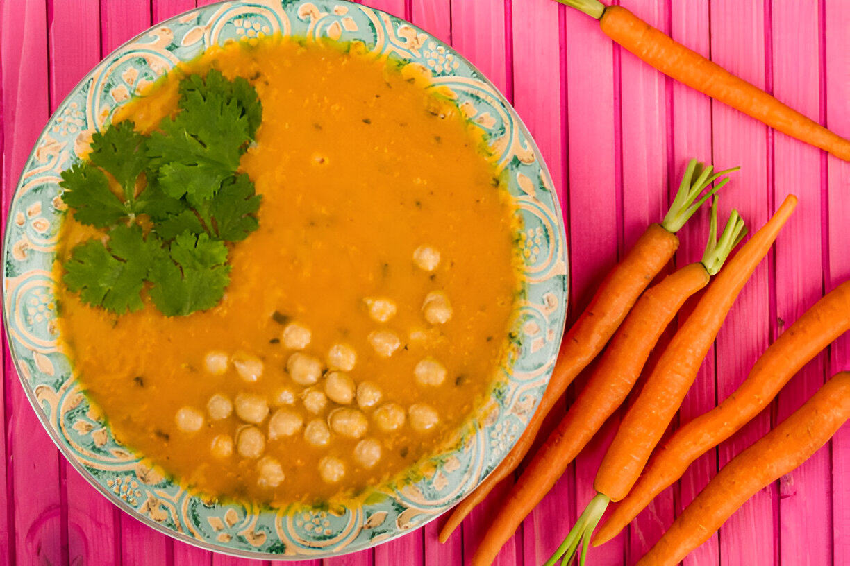 A plate of orange soup garnished with fresh cilantro, accompanied by fresh carrots on a pink wooden surface.
