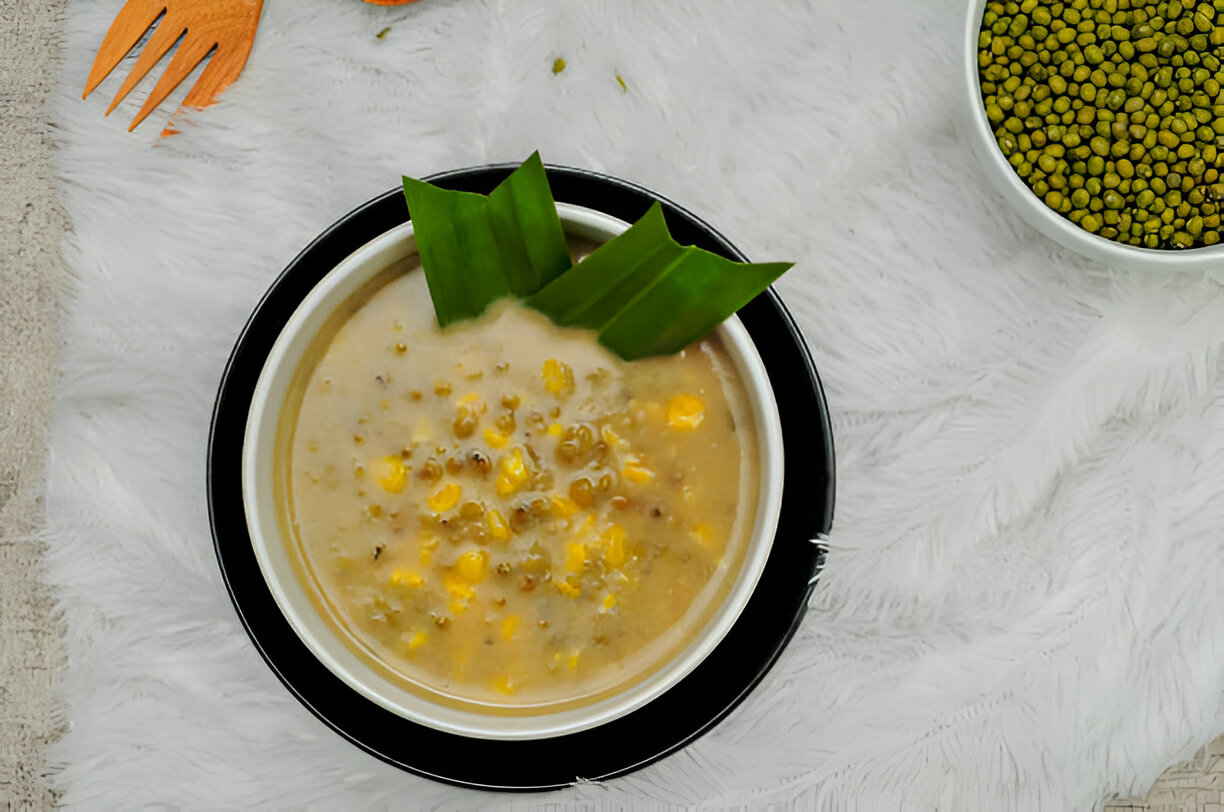A bowl of creamy soup with corn and mung beans, garnished with pandan leaves, placed on a white fluffy surface.