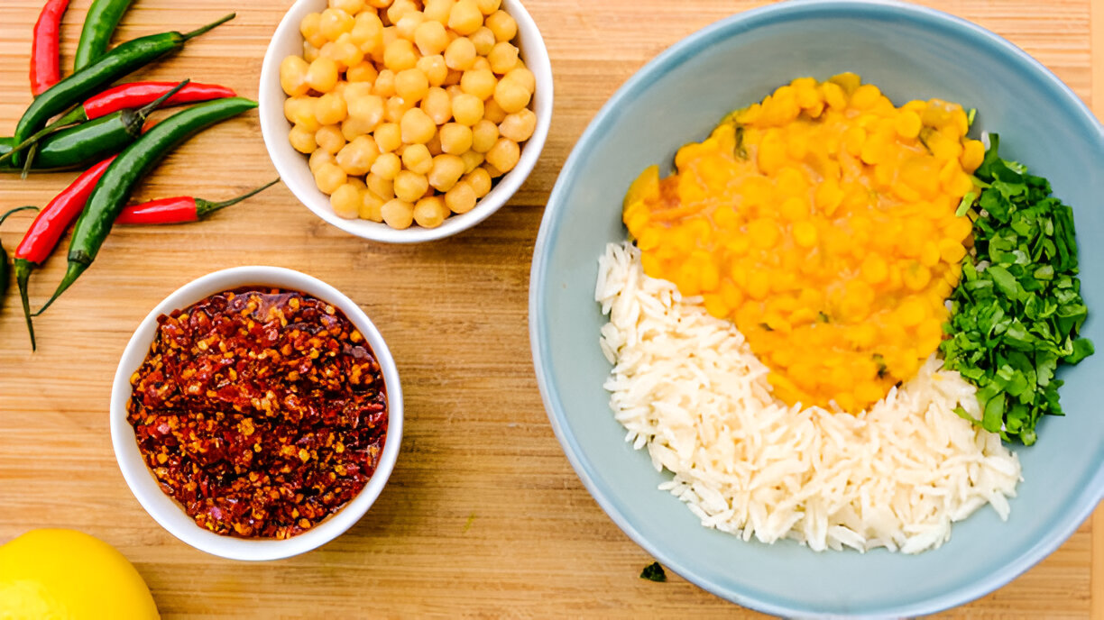 A colorful assortment of food ingredients on a wooden surface, including chickpeas, rice, herbs, chilies, and a spicy mixture.
