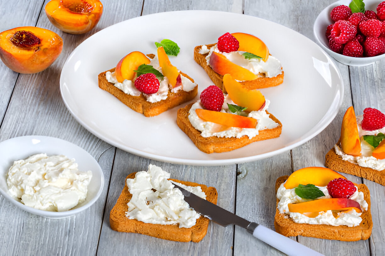 A plate of toast topped with cream cheese, sliced peaches, raspberries, and mint leaves, surrounded by fresh peaches and a small bowl of cream cheese.