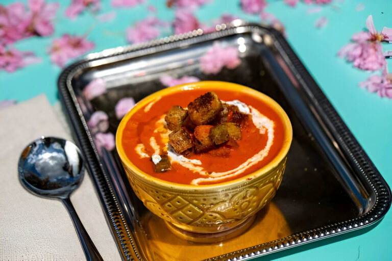 A golden bowl of rich red soup topped with crunchy croutons and a swirl of cream, served on a silver tray.