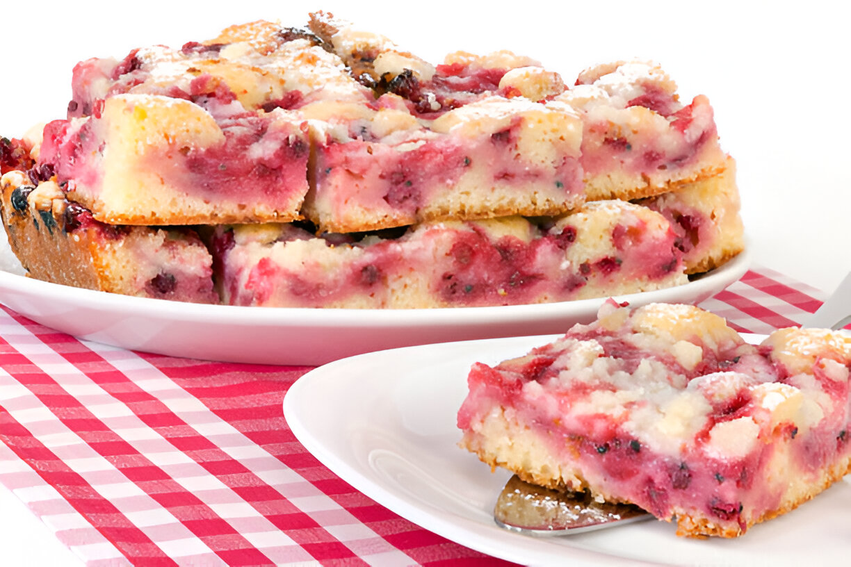A slice of colorful berry cake served on a white plate next to a stack of berry cake pieces.