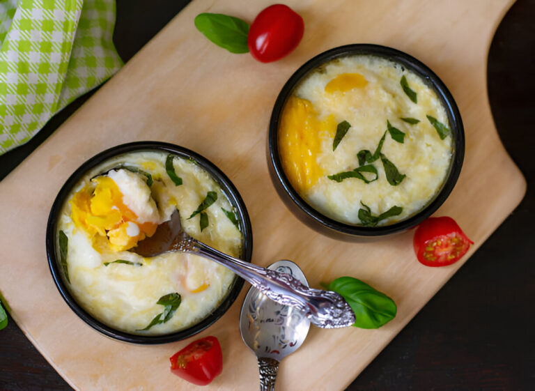 Two bowls of creamy egg dish with herbs on a wooden cutting board, surrounded by cherry tomatoes and a spoon.