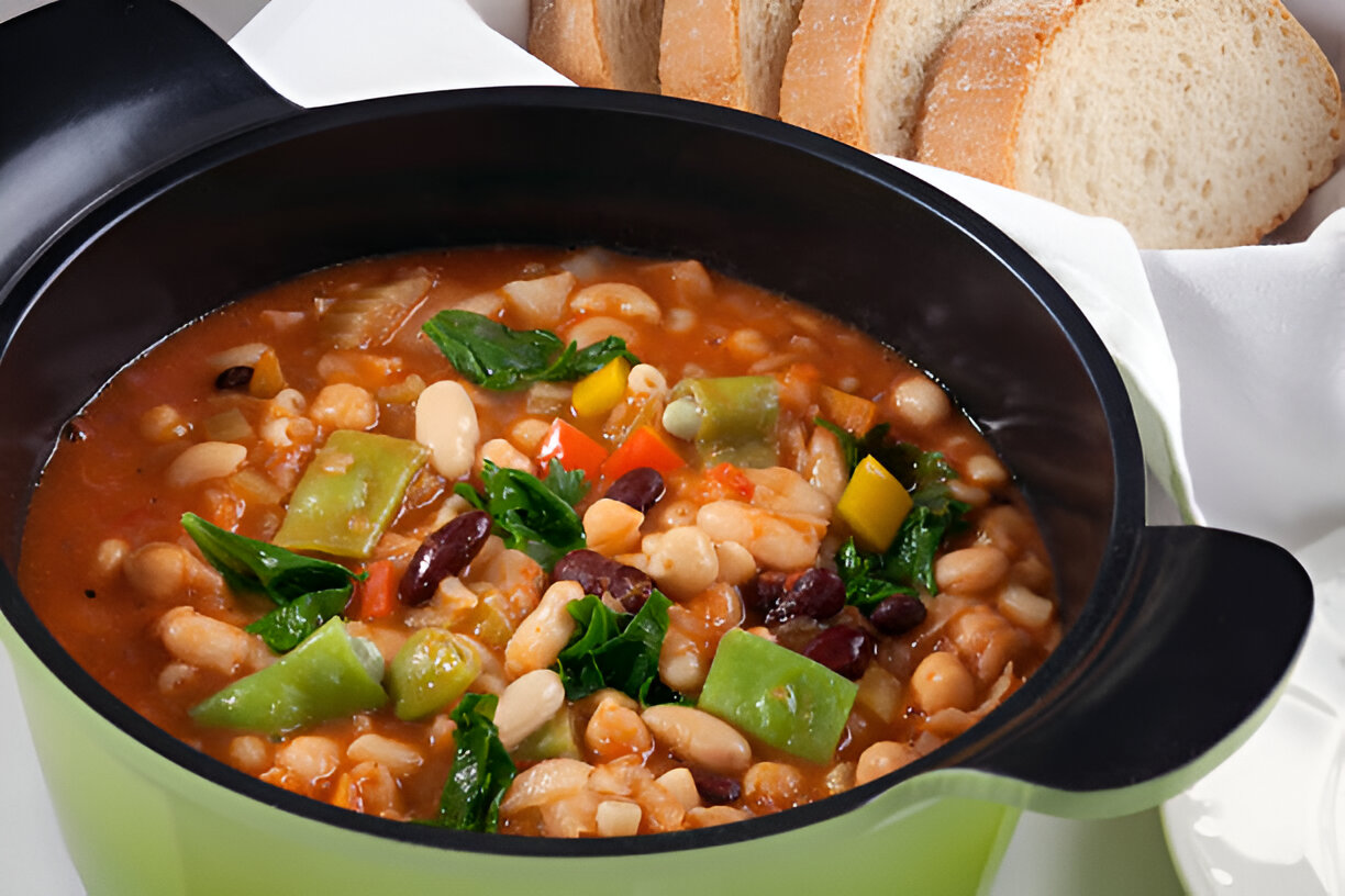 A bowl of hearty vegetable and bean soup with a variety of colorful vegetables, including greens, red and yellow peppers, and beans, next to slices of bread.