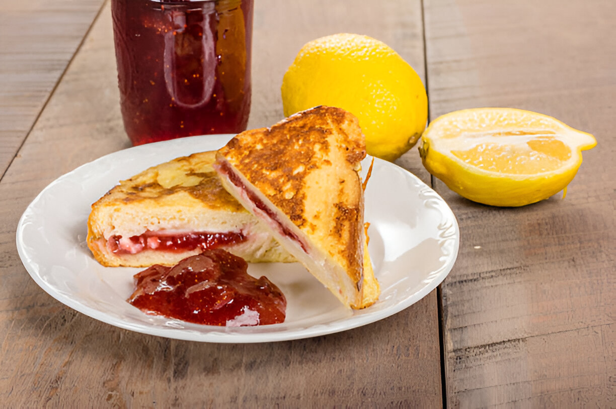 A slice of grilled French toast filled with strawberry jam on a white plate, accompanied by a small dollop of jam and fresh lemons.