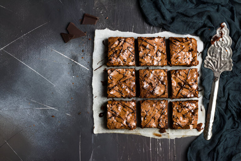 A freshly baked tray of brownies cut into squares, drizzled with chocolate, on parchment paper with a decorative serving spatula beside it.