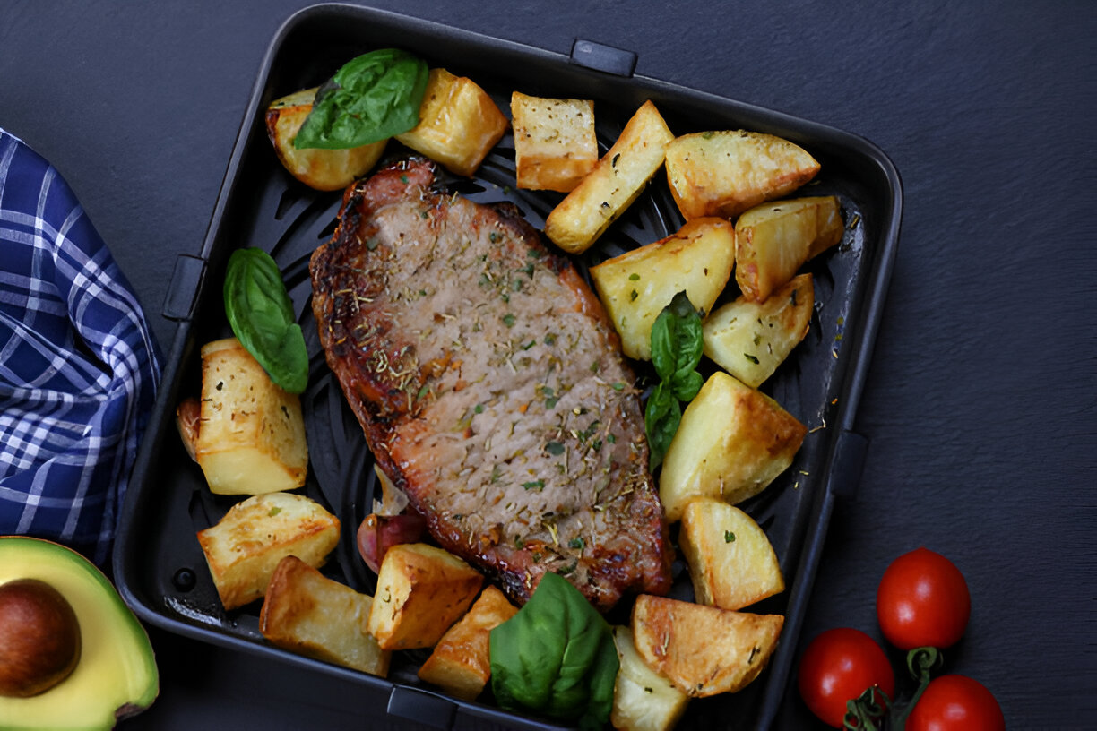 A roasted steak served with golden crispy potatoes and fresh basil on a black tray, alongside an avocado and cherry tomatoes.