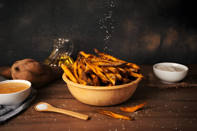 A wooden bowl filled with crispy sweet potato fries, sprinkled with salt, accompanied by sauces, a whole sweet potato, and a bottle of oil on a wooden surface.