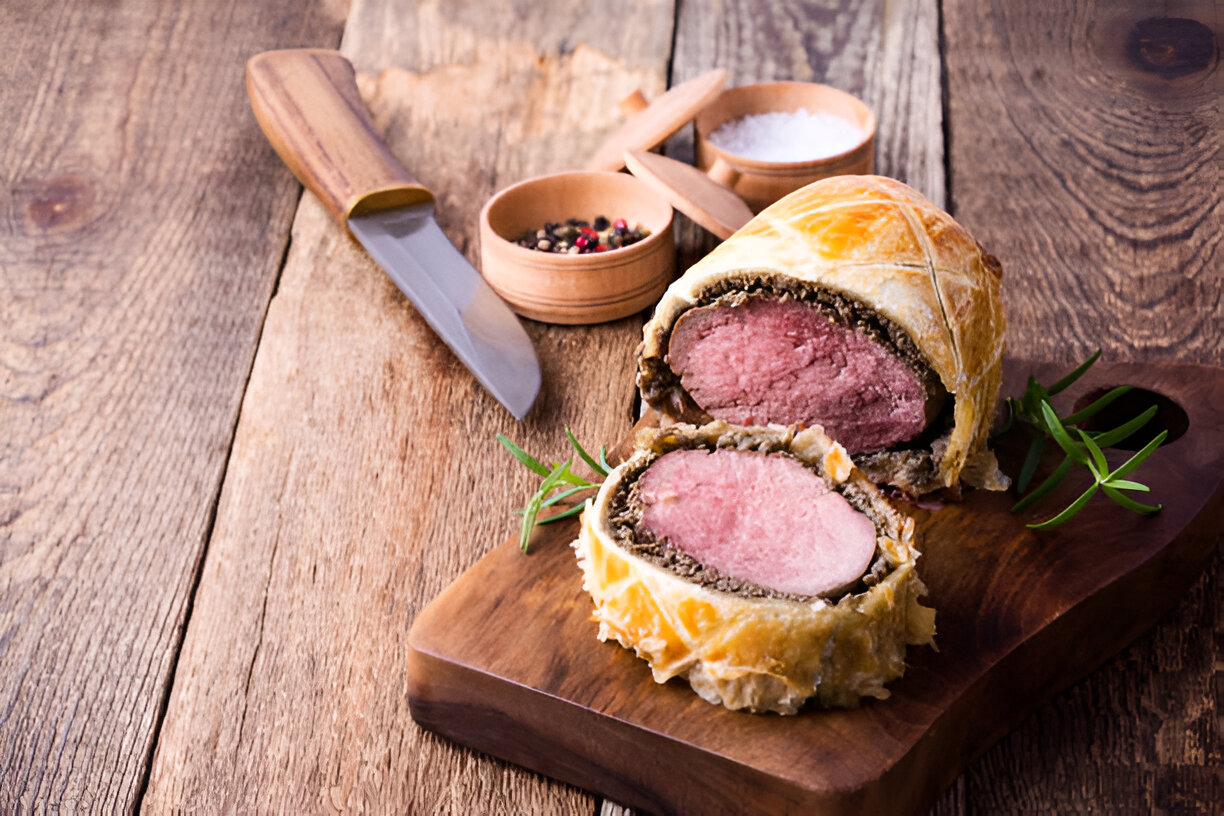 A beautifully plated portion of Beef Wellington, showcasing the tender pink meat encased in golden pastry, accompanied by a knife and small bowls of salt and peppercorns.