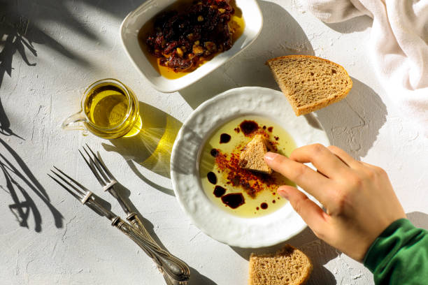 A hand dipping a piece of bread into a bowl of olive oil and spices, with a side dish of olives and a glass of olive oil on a white table.