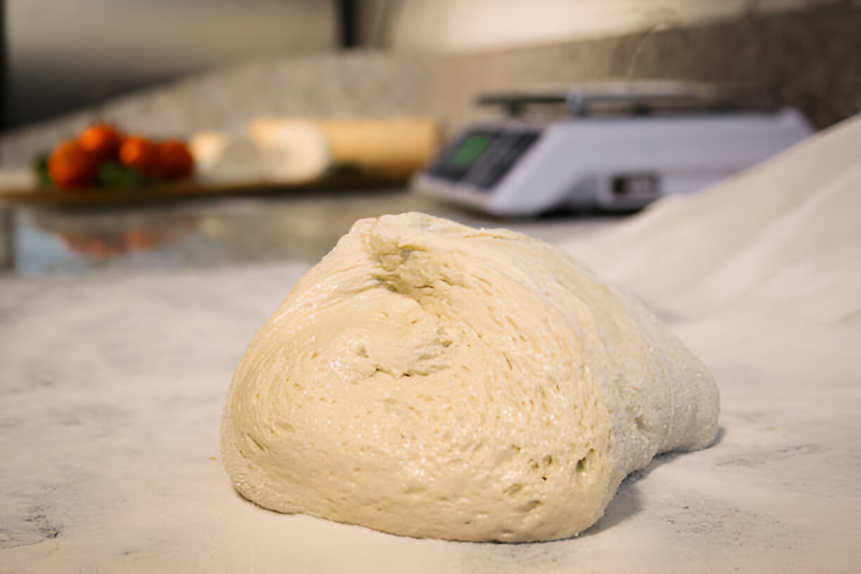 A close-up of a lump of pizza dough on a marble surface with flour scattered around, and a blurred background featuring tomatoes and cheese.