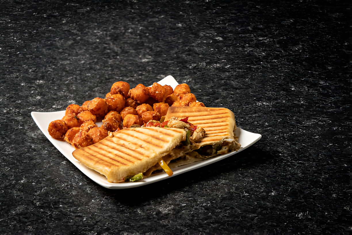 A white plate with a grilled sandwich and golden tater tots on a black marble surface.