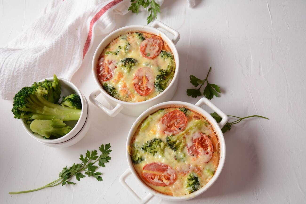 Two white bowls of baked broccoli and tomato dish next to a bowl of fresh broccoli on a light surface.