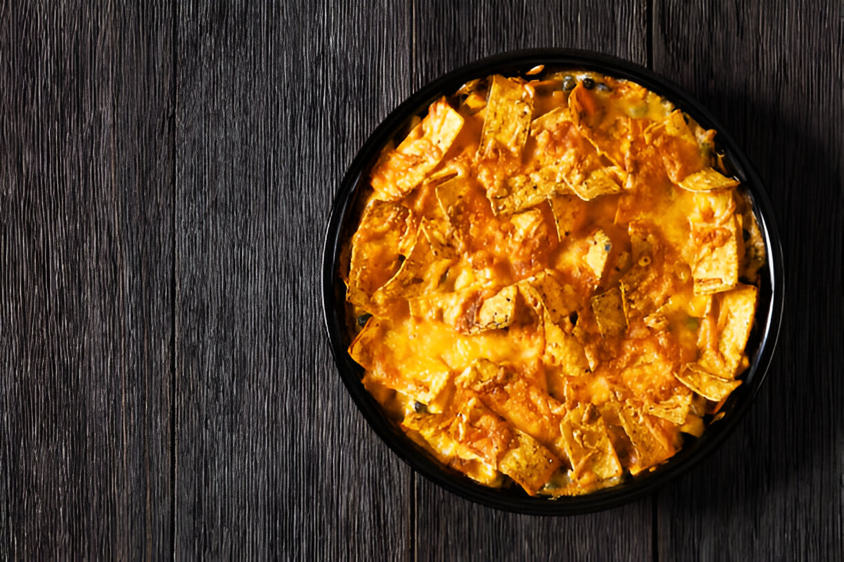 A delicious baked dish topped with golden crunchy tortilla chips and melted cheese in a black bowl on a wooden surface.