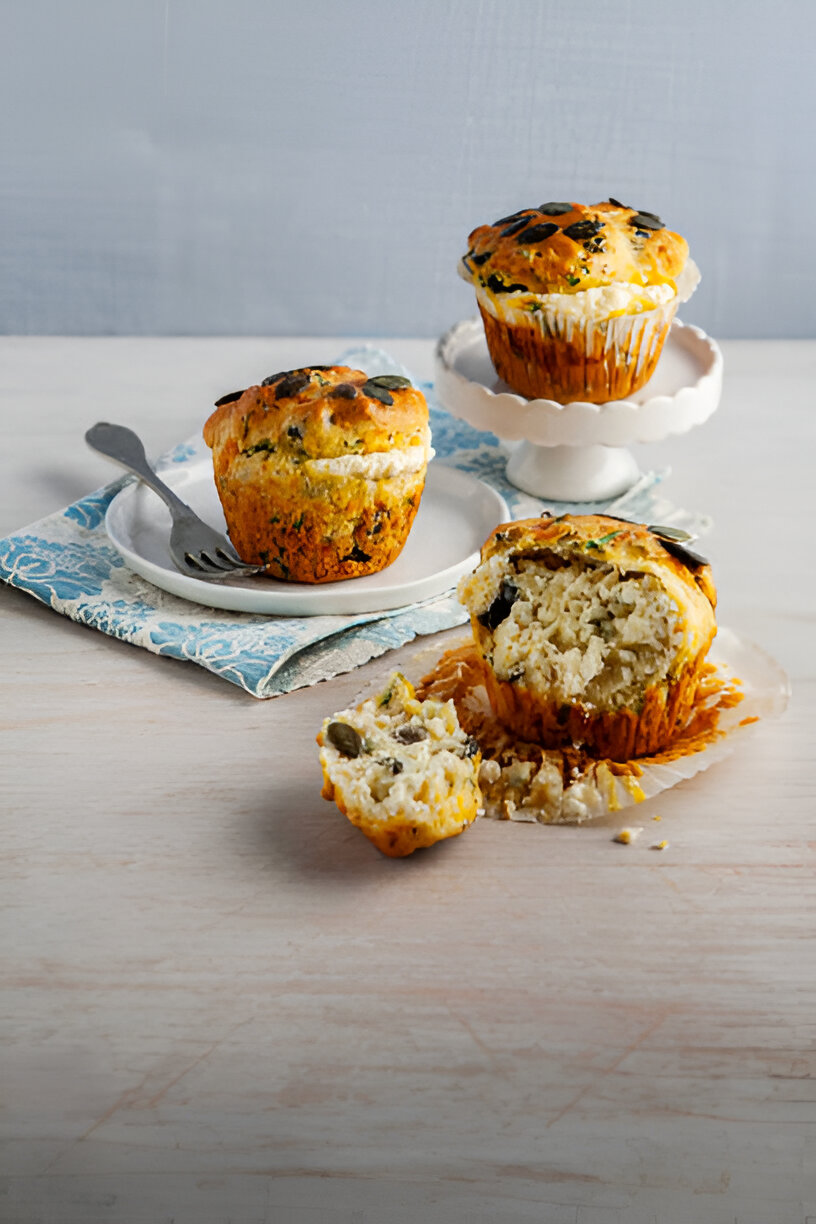 Three savory muffins displayed on a white plate and a cake stand, with one muffin partially broken apart showing its filling.