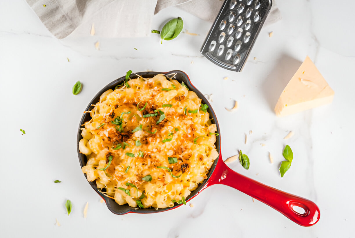 A serving of creamy macaroni and cheese in a small cast iron skillet, garnished with basil leaves and surrounded by cheese and a grater.