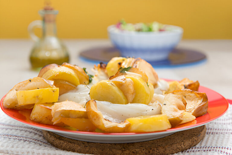 A colorful plate of roasted potatoes and fish garnished with herbs, served on a red plate with a blurred background of a salad bowl.